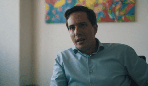 Stefan Duhr looking left, wearing blue buttoned shirt, blurry background with painting