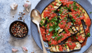 Eggplants with red sauce and parsley garnish on a gray plate next to garlic and seasoning